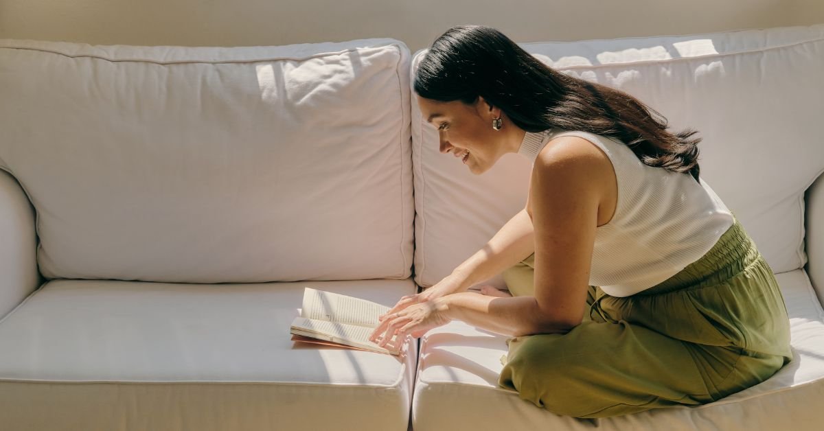 A woman sitting cross-legged on a couch, wearing a white top and loose green pants, intently reading a book in a relaxed, cozy setting.