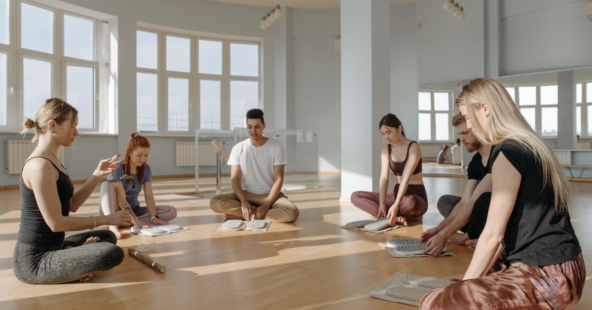 A group of six people sitting cross-legged in a circle, focused and leaning over colorful acupressure mats during a wellness or mindfulness session.