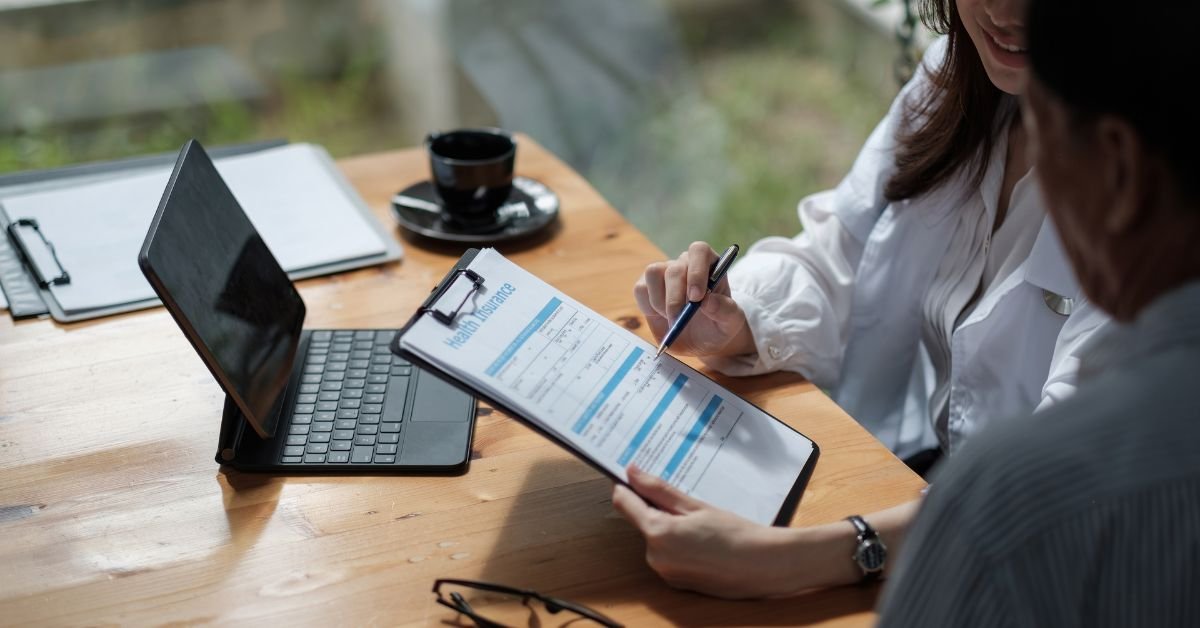 Woman practicing self-care with hands over heart, wearing chakra bracelets and a floral kimono. Two people are sitting at a table nearby with a tablet; one is holding a health questionnaire — symbolizing wellness coaching and mindful health assessment.