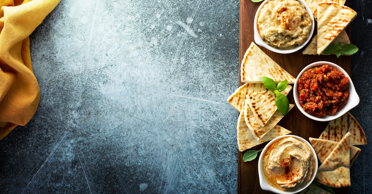 Top view of assorted vegan spreads and bread on a dark background, arranged with rustic kitchen elements.